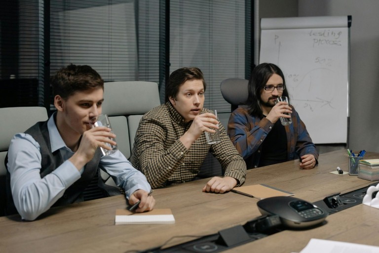 Three men in a meeting room drinking water during a business discussion.