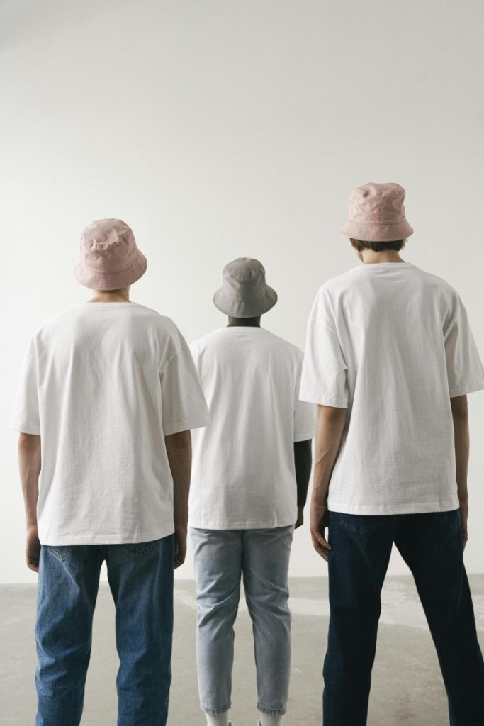 Three men in bucket hats and white shirts, photographed from behind in a minimalist setting.